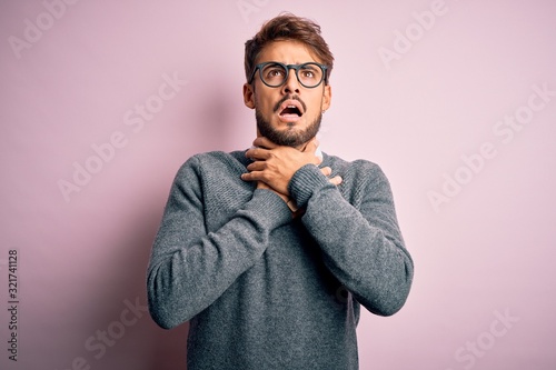 Young handsome man with beard wearing glasses and sweater standing over pink background shouting and suffocate because painful strangle. Health problem. Asphyxiate and suicide concept.