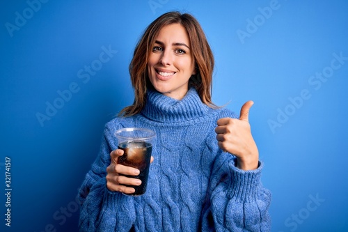 Young beautiful brunette woman drinking glass with cola refreshment over blue background happy with big smile doing ok sign  thumb up with fingers  excellent sign