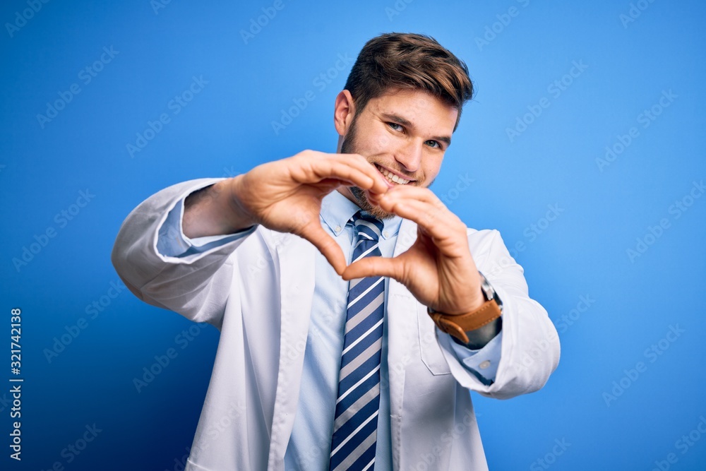 Εκτύπωση καμβά Young blond therapist man with beard and blue eyes wearing coat and tie over background smiling in love doing heart symbol shape with hands