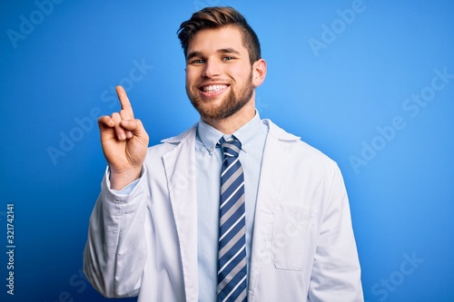 Young blond therapist man with beard and blue eyes wearing coat and tie over background showing and pointing up with finger number one while smiling confident and happy.