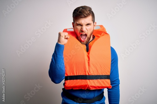 Young blond tourist man with beard and blue eyes wearing lifejacket over white background angry and mad raising fist frustrated and furious while shouting with anger. Rage and aggressive concept. © Krakenimages.com