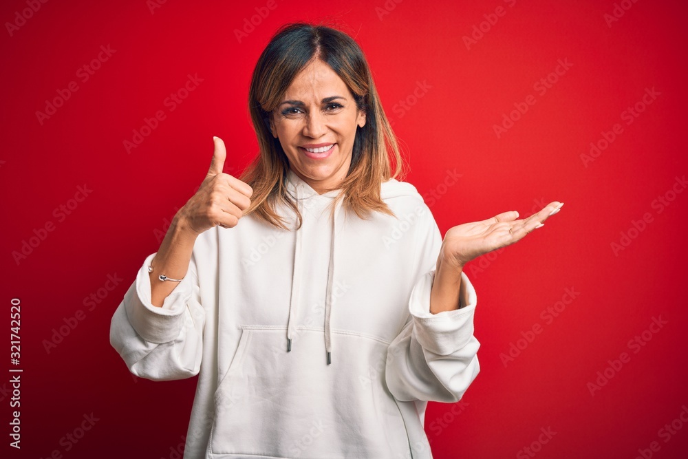 Middle age beautiful sportswoman wearing casual sweatshirt over isolated red background Showing palm hand and doing ok gesture with thumbs up, smiling happy and cheerful Middle age beautiful sportswoman wearing casual sweatshirt over isolated red background Showing palm hand and doing ok gesture with thumbs up, smiling happy and cheerful