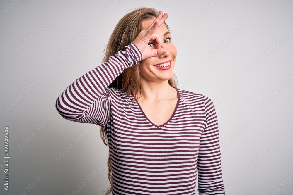 Young beautiful blonde woman wearing casual striped t-shirt over isolated white background doing ok gesture with hand smiling, eye looking through fingers with happy face. Young beautiful blonde woman wearing casual striped t-shirt over isolated white background doing ok gesture with hand smiling, eye looking through fingers with happy face.