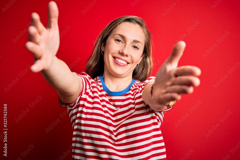 Young beautiful blonde woman wearing casual striped t-shirt over isolated red background looking at the camera smiling with open arms for hug. Cheerful expression embracing happiness. Young beautiful blonde woman wearing casual striped t-shirt over isolated red background looking at the camera smiling with open arms for hug. Cheerful expression embracing happiness.