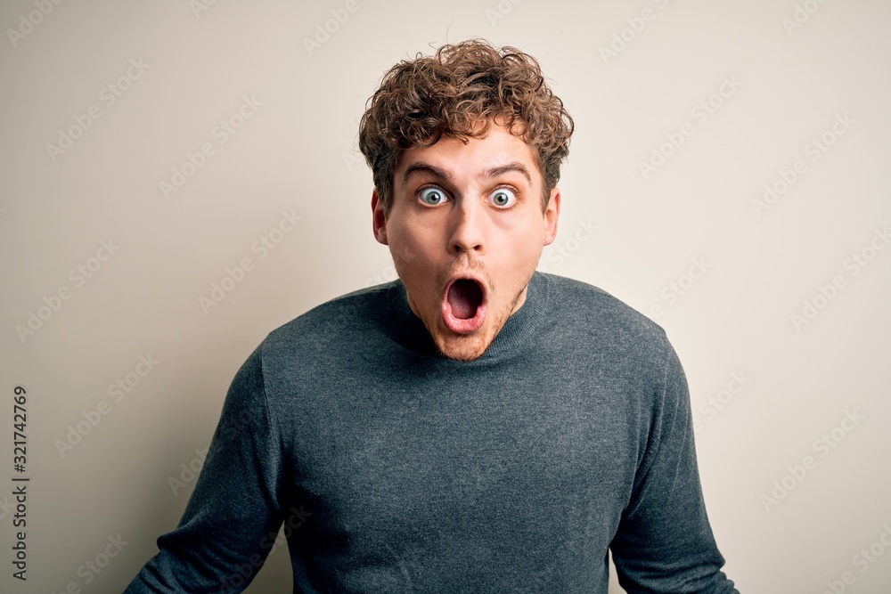 Young blond handsome man with curly hair wearing casual sweater over white background afraid and shocked with surprise expression, fear and excited face. Young blond handsome man with curly hair wearing casual sweater over white background afraid and shocked with surprise expression, fear and excited face.