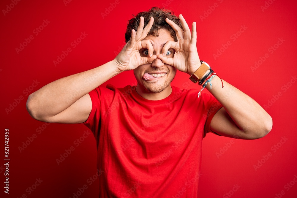 Young blond handsome man with curly hair wearing casual t-shirt over red background doing ok gesture like binoculars sticking tongue out, eyes looking through fingers. Crazy expression.