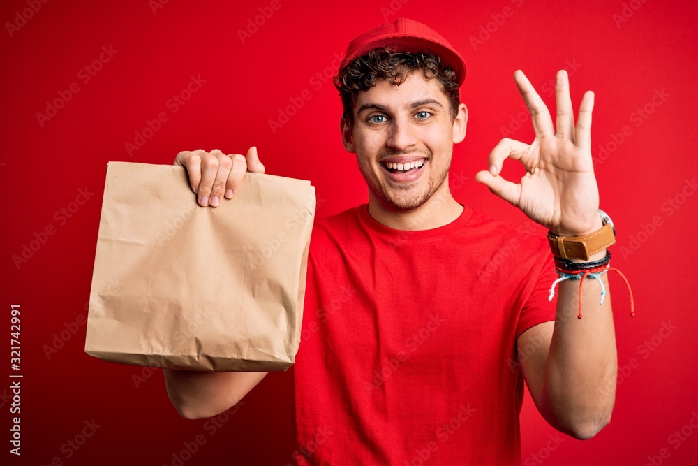 Young blond delivery man with curly hair wearing cap holding paper bag with food doing ok sign with fingers, excellent symbol Young blond delivery man with curly hair wearing cap holding paper bag with food doing ok sign with fingers, excellent symbol
