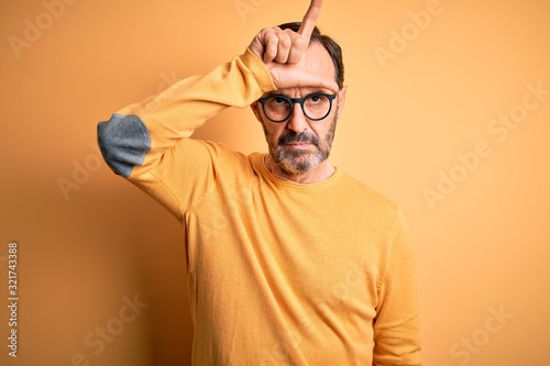 Middle age hoary man wearing casual sweater and glasses over isolated yellow background making fun of people with fingers on forehead doing loser gesture mocking and insulting.