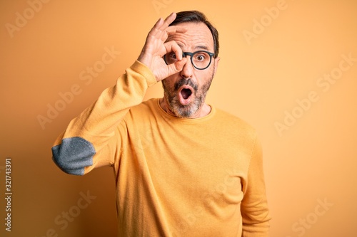 Middle age hoary man wearing casual sweater and glasses over isolated yellow background doing ok gesture shocked with surprised face, eye looking through fingers. Unbelieving expression.