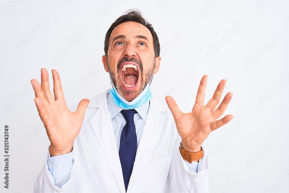 Middle age scientist man wearing coat and medical mask over isolated white background crazy and mad shouting and yelling with aggressive expression and arms raised. Frustration concept. Middle age scientist man wearing coat and medical mask over isolated white background crazy and mad shouting and yelling with aggressive expression and arms raised. Frustration concept.