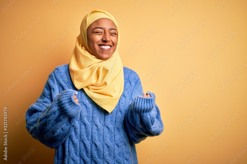 Young African American afro woman wearing muslim hijab over isolated yellow background excited for success with arms raised and eyes closed celebrating victory smiling. Winner concept. Young African American afro woman wearing muslim hijab over isolated yellow background excited for success with arms raised and eyes closed celebrating victory smiling. Winner concept.