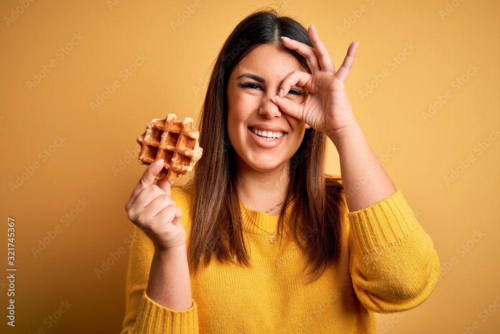 Young beautiful woman eating sweet waffle pastry over yellow background with happy face smiling doing ok sign with hand on eye looking through fingers Young beautiful woman eating sweet waffle pastry over yellow background with happy face smiling doing ok sign with hand on eye looking through fingers