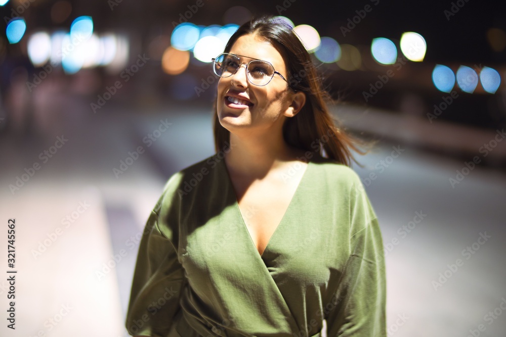 Young beautiful girl smiling happy and confident walking at the promenade, standing with a smile on face around night lights bokeh
