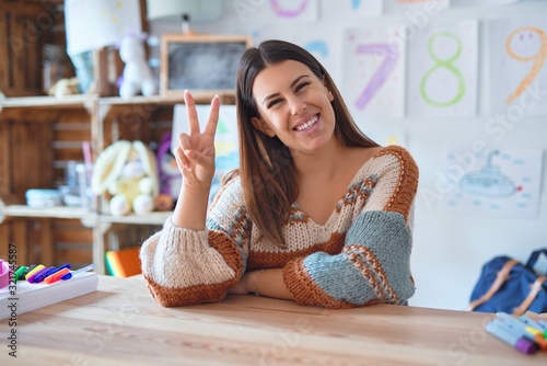 Young beautiful teacher woman wearing sweater and glasses sitting on desk at kindergarten smiling with happy face winking at the camera doing victory sign. Number two.