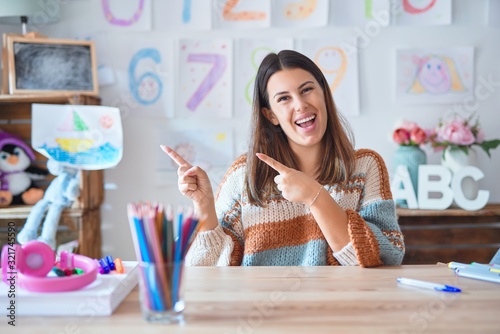 Young beautiful teacher woman wearing sweater and glasses sitting on desk at kindergarten smiling and looking at the camera pointing with two hands and fingers to the side.
