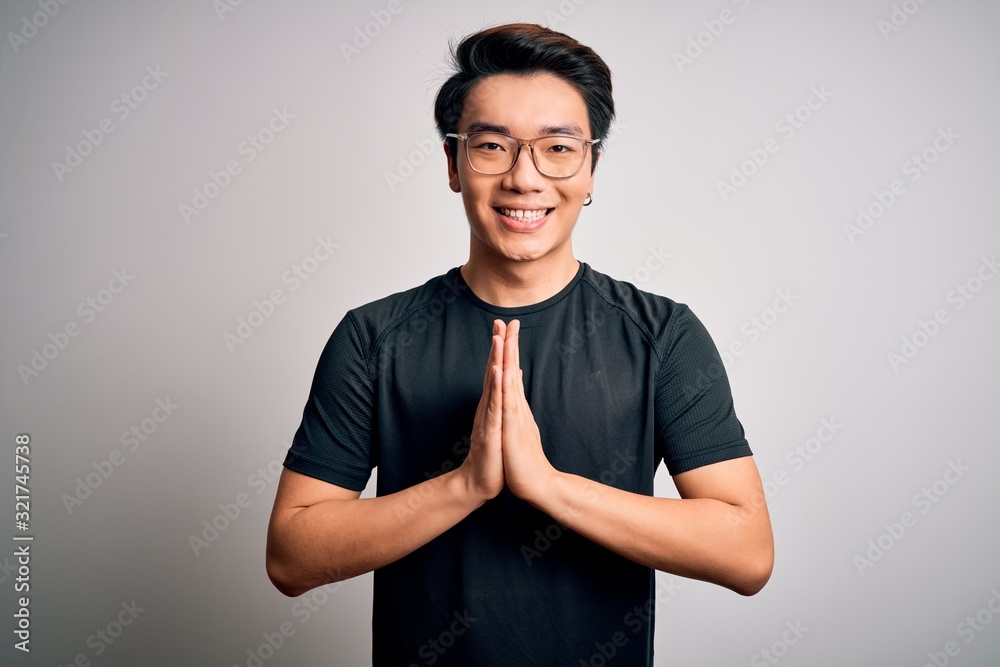 Young handsome chinese man wearing black t-shirt and glasses over white background praying with hands together asking for forgiveness smiling confident. Young handsome chinese man wearing black t-shirt and glasses over white background praying with hands together asking for forgiveness smiling confident.