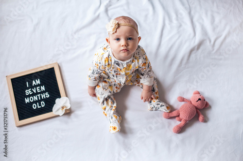 7 Seven months old baby girl sitting on white background with letter board and teddy bear. Flat lay composition.