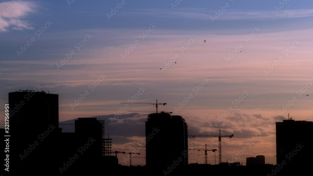 Aerial shot (Time Lapse 4K) of city silhouette from dusk to night with flying clouds and sky color changes. In the background working cranes and skyscraper building in the process of construction. Aerial shot (Time Lapse 4K) of city silhouette from dusk to night with flying clouds and sky color changes. In the background working cranes and skyscraper building in the process of construction.