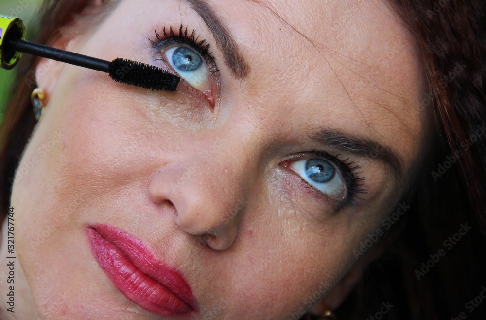 brunette woman blue-eyed applying mascara on her long eyelashes, close-up. look up. everyday makeup, red lipstick on the lips