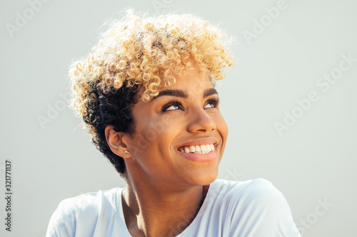 hispanic latin woman with afro blonde haircut dressed in white top looking up with charming shy smile in studio white background