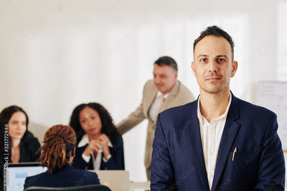 Portrait of smiling handsome businessman looking at camera when his coworker working on new creative project in background Portrait of smiling handsome businessman looking at camera when his coworker working on new creative project in background