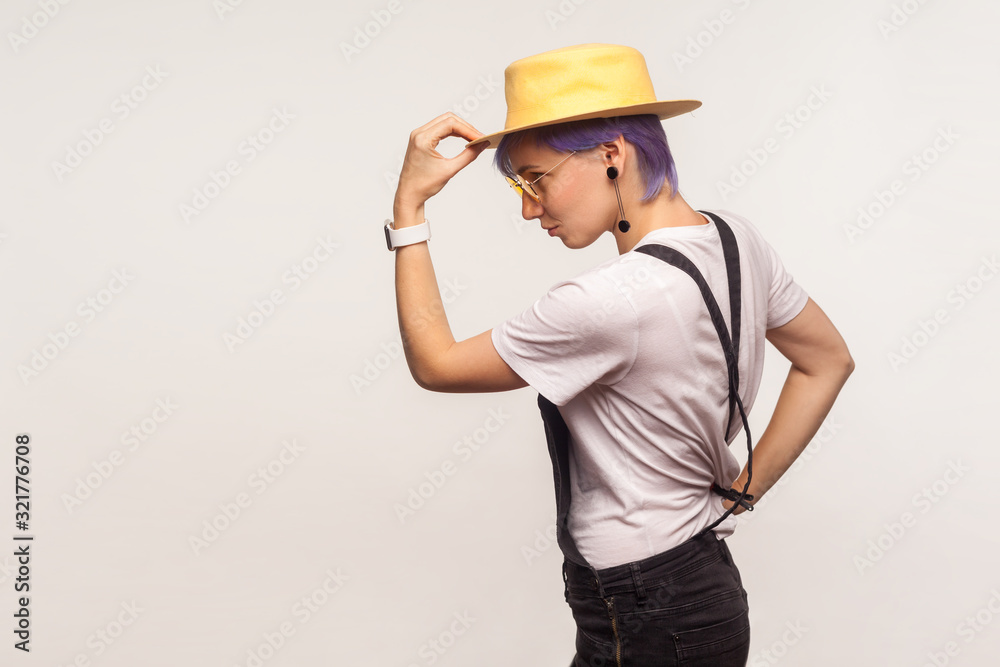 Side view portrait of elegant stylish hipster girl with violet hair and in overalls holding hat on head, looking at empty space for your advertisement. isolated on white background, studio shot