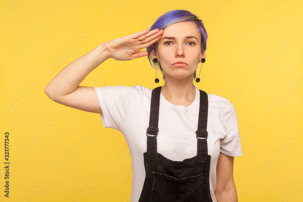 Yes sir! Portrait of serious obedient hipster girl with violet short hair in denim overalls keeping hand near temple and saluting as soldier, listening to commander. yellow background, studio shot