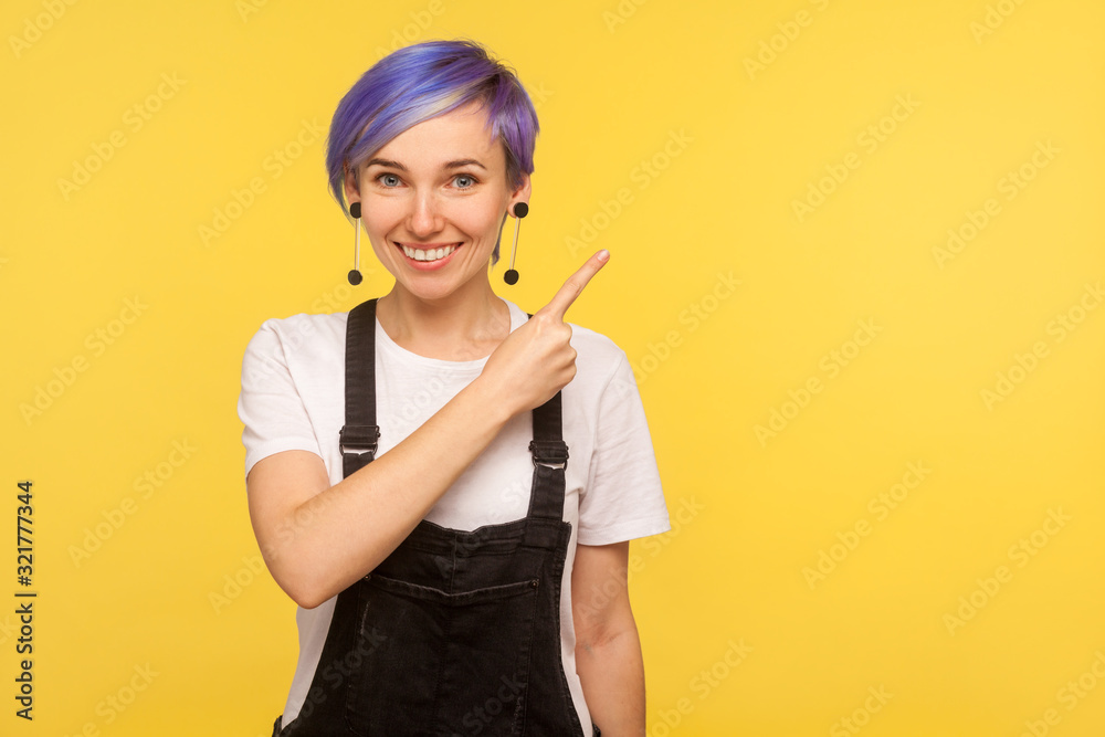 Portrait of fashionable positive smiling hipster girl with violet short hair in denim overalls pointing to the side freespace for advertising, copy space. isolated on yellow background, studio shot Portrait of fashionable positive smiling hipster girl with violet short hair in denim overalls pointing to the side freespace for advertising, copy space. isolated on yellow background, studio shot
