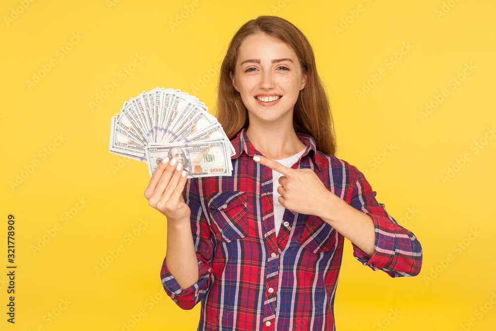 Look at my income! Portrait of delighted girl in checkered shirt pointing at dollars and smiling excitedly, boasting of profit, showing cash to camera. indoor studio shot isolated on yellow background Look at my income! Portrait of delighted girl in checkered shirt pointing at dollars and smiling excitedly, boasting of profit, showing cash to camera. indoor studio shot isolated on yellow background
