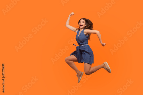 Fototapeta Naklejka Na Ścianę i Meble -  Leap to success. Full length of excited energetic motivated brunette woman in denim dress running in air, hurrying to achieve goal, jumping rejoicing victory. studio shot isolated on orange background