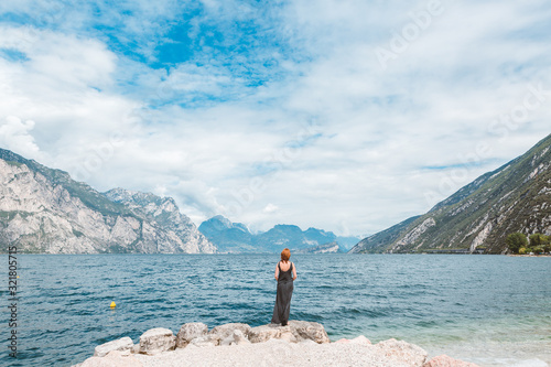 A woman in a long gray dress is standing on the rocky shore of Lake Garda. Italian landscape on a summer sunny day. photo