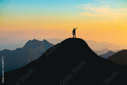 The silhouette of a man with a cheerful raise in his hand on the top of the mountain During sunset time The concept of effort and success photo