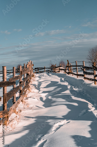 Winter season, landscape picture of a village road covered by snow at sunset, footpath in snow and old timber fence on the side of the road