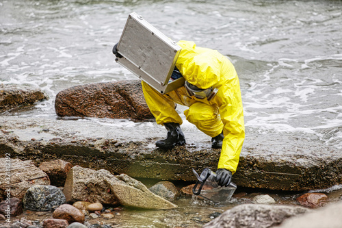 Tela specialist in protective suit taking sample of water to container on rocky shore