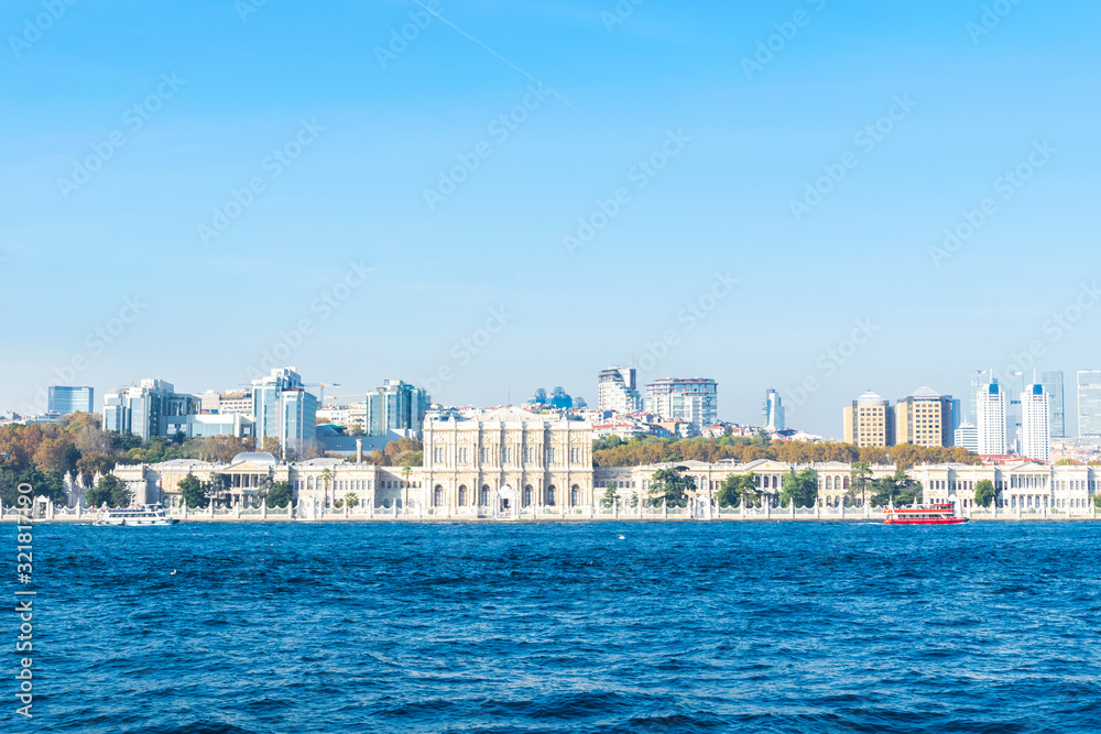 Dolmabahce palace building view from seaside with red and white tourist ferry around and Besiktas district area in the background. Istanbul, Turkey. Historical buildings and tourism industry in Turkey