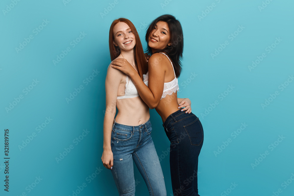 two best friends in stulish jeans and white cotton bras having fun in the studio, entertainment, clothes, fashion, style, isolated blue bacakground, studio shot, asian girl hugging her ginger friend two best friends in stulish jeans and white cotton bras having fun in the studio, entertainment, clothes, fashion, style, isolated blue bacakground, studio shot, asian girl hugging her ginger friend