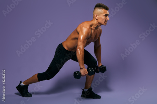 young motivated atrong man doing loungers with dumbbells, full length side view photo, isolated blue background, studio shot