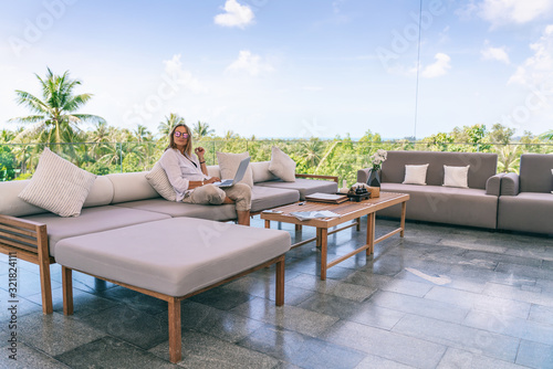 Wallpaper Mural Attractive woman 40 years old in a white shirt sitting on a gray sofa working on a laptop on the terrace overlooking the green jungle on a bright sunny day Torontodigital.ca