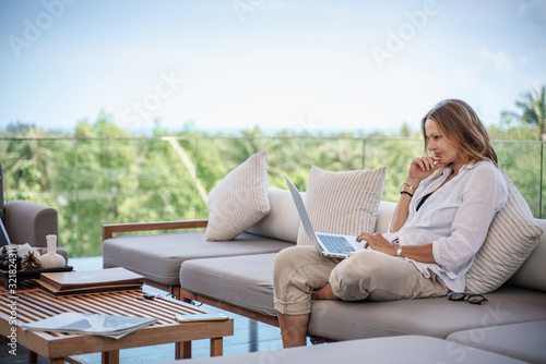Attractive woman 40 years old in a white shirt sitting on a gray sofa working on a laptop on the terrace overlooking the green jungle on a bright sunny day