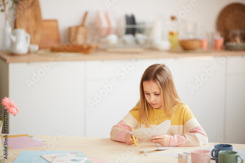 Wide angle portrait of cute girl making holiday card for Mothers day or Valentines day while sitting at table in cozy home interior, copy space