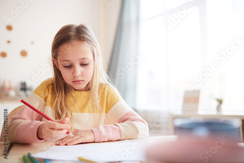 Warm toned portrait of cute little girl drawing pictures or doing homework while sitting at table in home interior, copy space