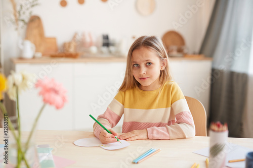 Warm toned portrait of cute little girl looking at camera and smiling while drawing pictures or doing homework while sitting at table in home interior, copy space
