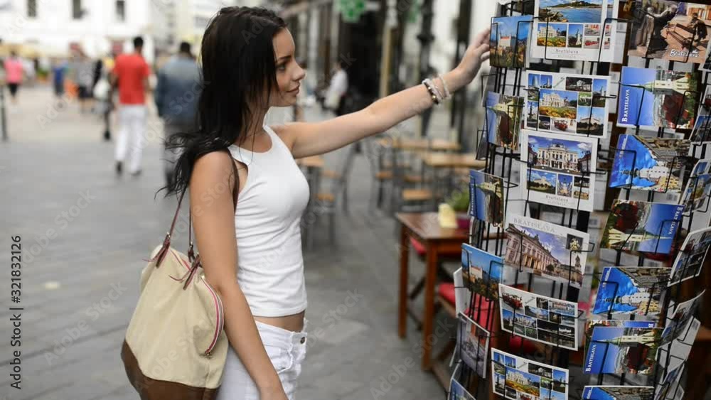 Teen black haired woman in white clothing is standing on the street, looking at postcard stand and choosing some postcard in historical part of Bratislava city.