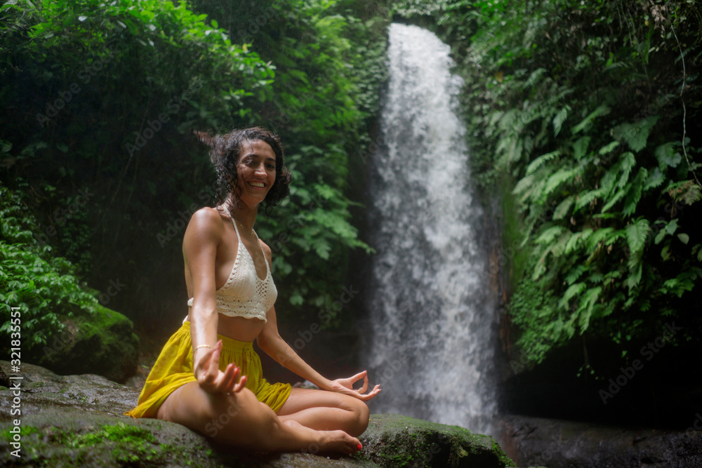 Fototapeta premium outdoors portrait of young attractive and happy hipster girl doing yoga at beautiful tropical waterfall meditating enjoying freedom and¡ nature in wellness and zen lifestyle