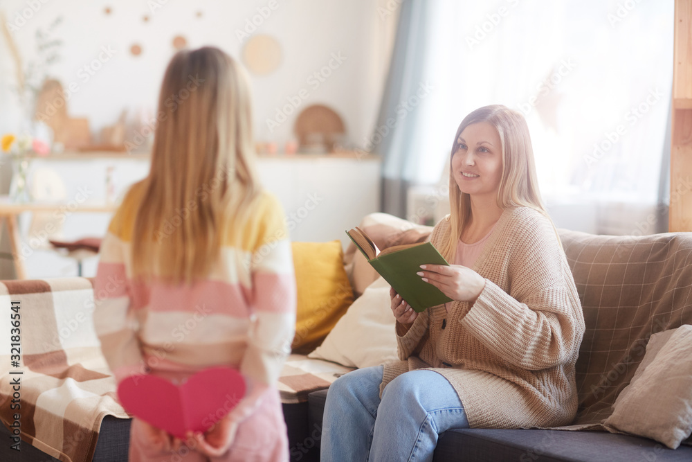 Warm-toned portrait of happy modern mom looking at daughter surprising her on mothers day while reading book on couch in cozy home interior, copy space Warm-toned portrait of happy modern mom looking at daughter surprising her on mothers day while reading book on couch in cozy home interior, copy space
