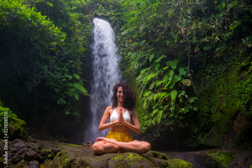 outdoors portrait of young attractive and happy hipster girl doing yoga at beautiful tropical waterfall meditating enjoying freedom and   nature in wellness and zen lifestyle