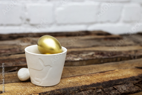 White pot with decorative bunny and Easter golden eggs on brown wooden table and white bricked background. Easter rabbit, eco decoration, spring. Copy space