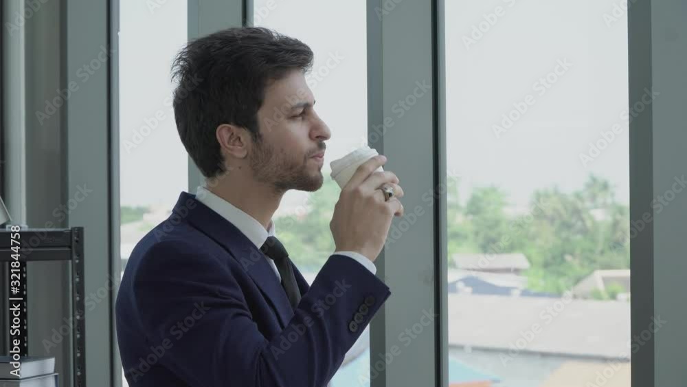 A young businessman in a black suit looking to the view outside the window, in his hand he held a cup of coffee, he drank the coffee and was thinking about something. A young businessman in a black suit looking to the view outside the window, in his hand he held a cup of coffee, he drank the coffee and was thinking about something.