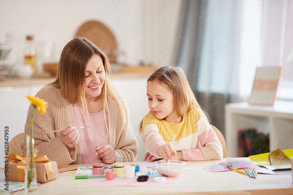 Warm-toned portrait of smiling mature mother painting pictures with cute little daughter while sitting at wooden table in cozy home interior Warm-toned portrait of smiling mature mother painting pictures with cute little daughter while sitting at wooden table in cozy home interior