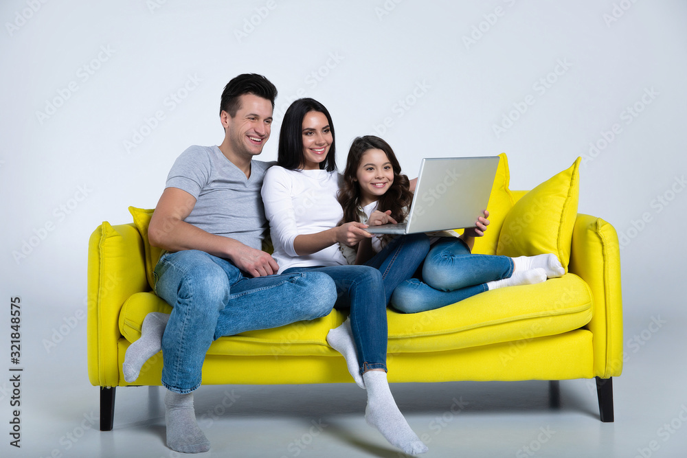 Family chilling on the sofa. A young family of three are watching films  from the laptop, relaxing together on a yellow couch in a living room.  Stock Photo | Adobe Stock